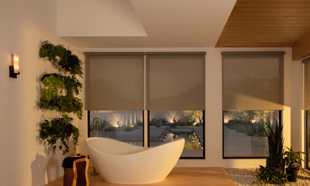 Zen bathroom with a white soaking tub and motorized shades overlooking a lit patio