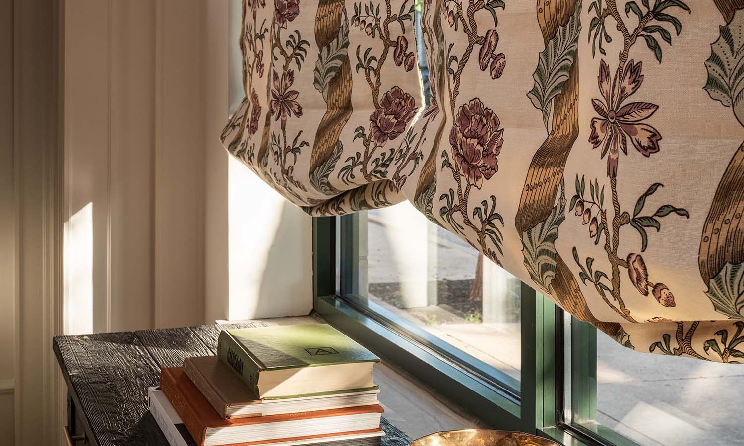 Patterned floral roman shades partially lowered over a window with books on the sill